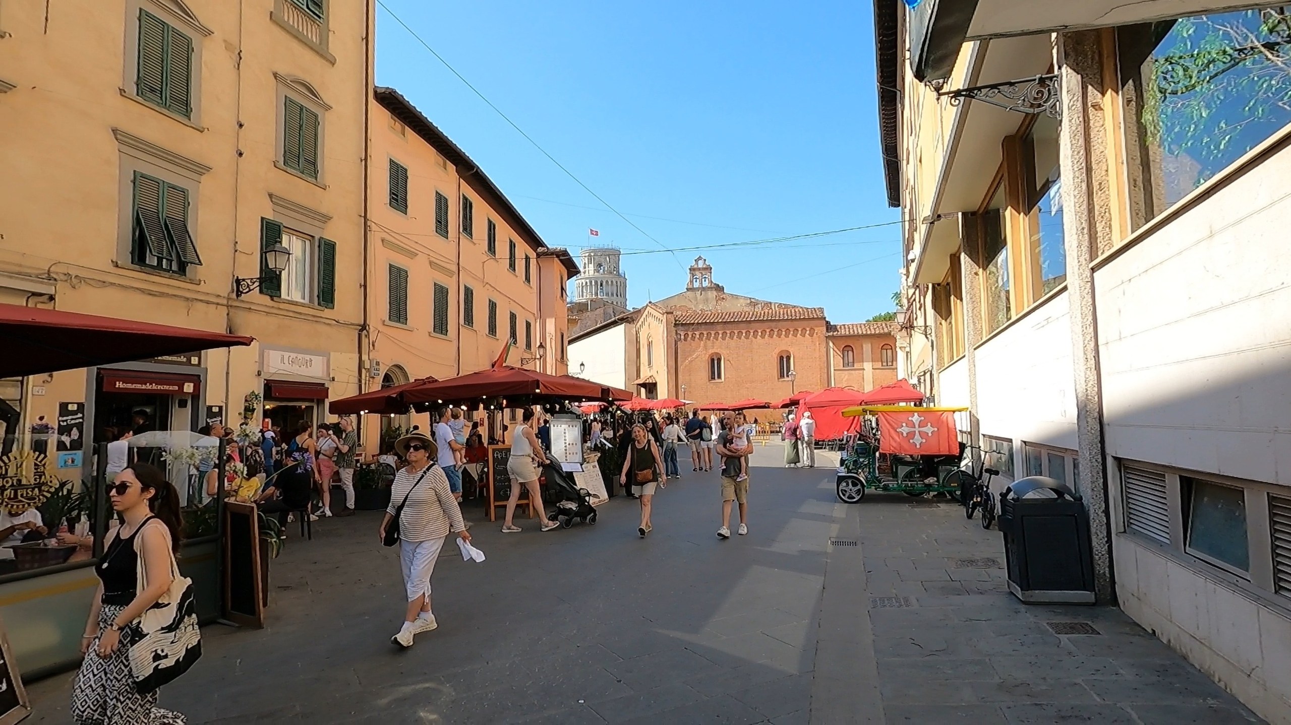 A street of cafes and market stalls near the Square of Miracles in Pisa, the Tower visible in the distance