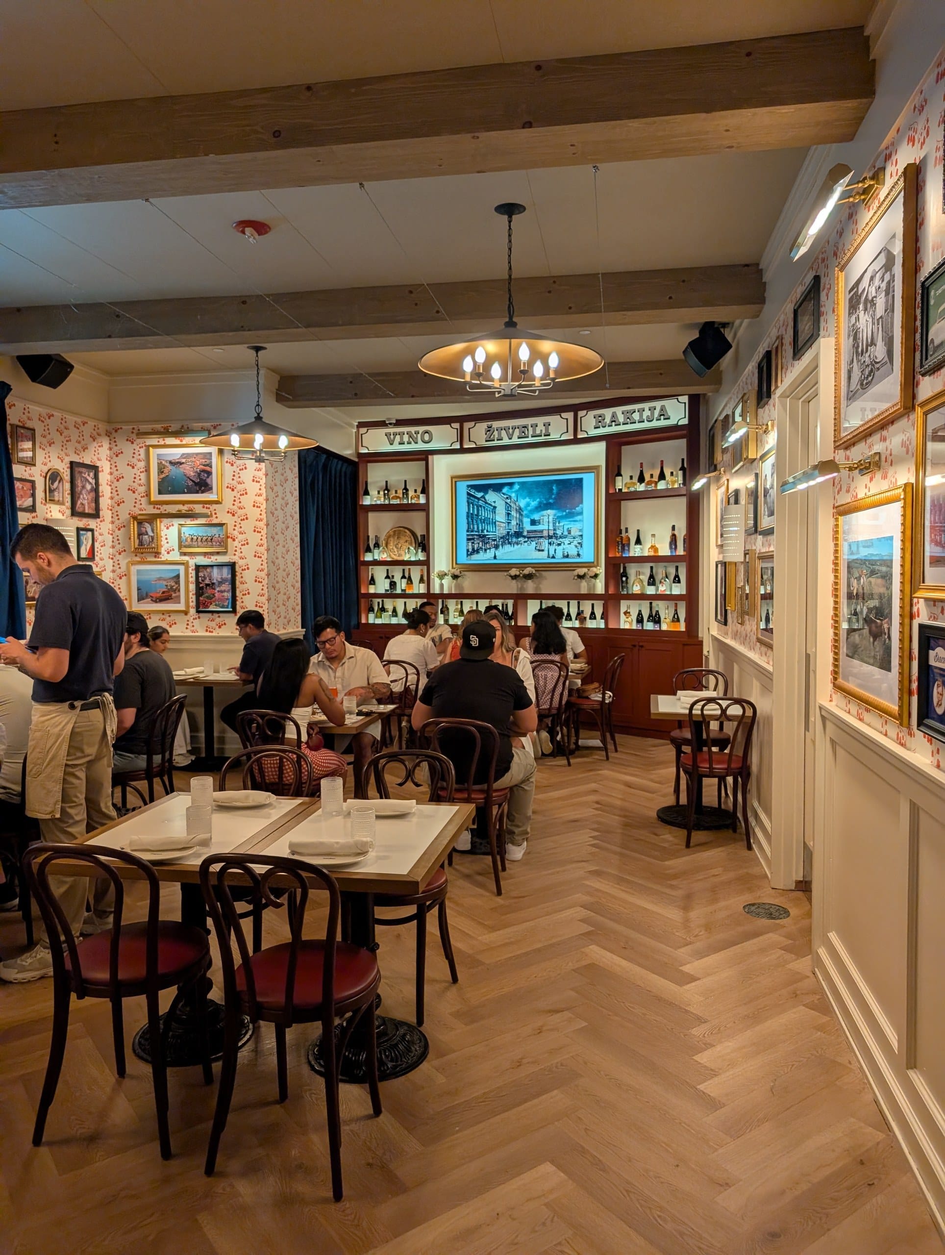 Tables fill the front room at midday under pendant lights, with the Balkan gallery wall running the length of the space.