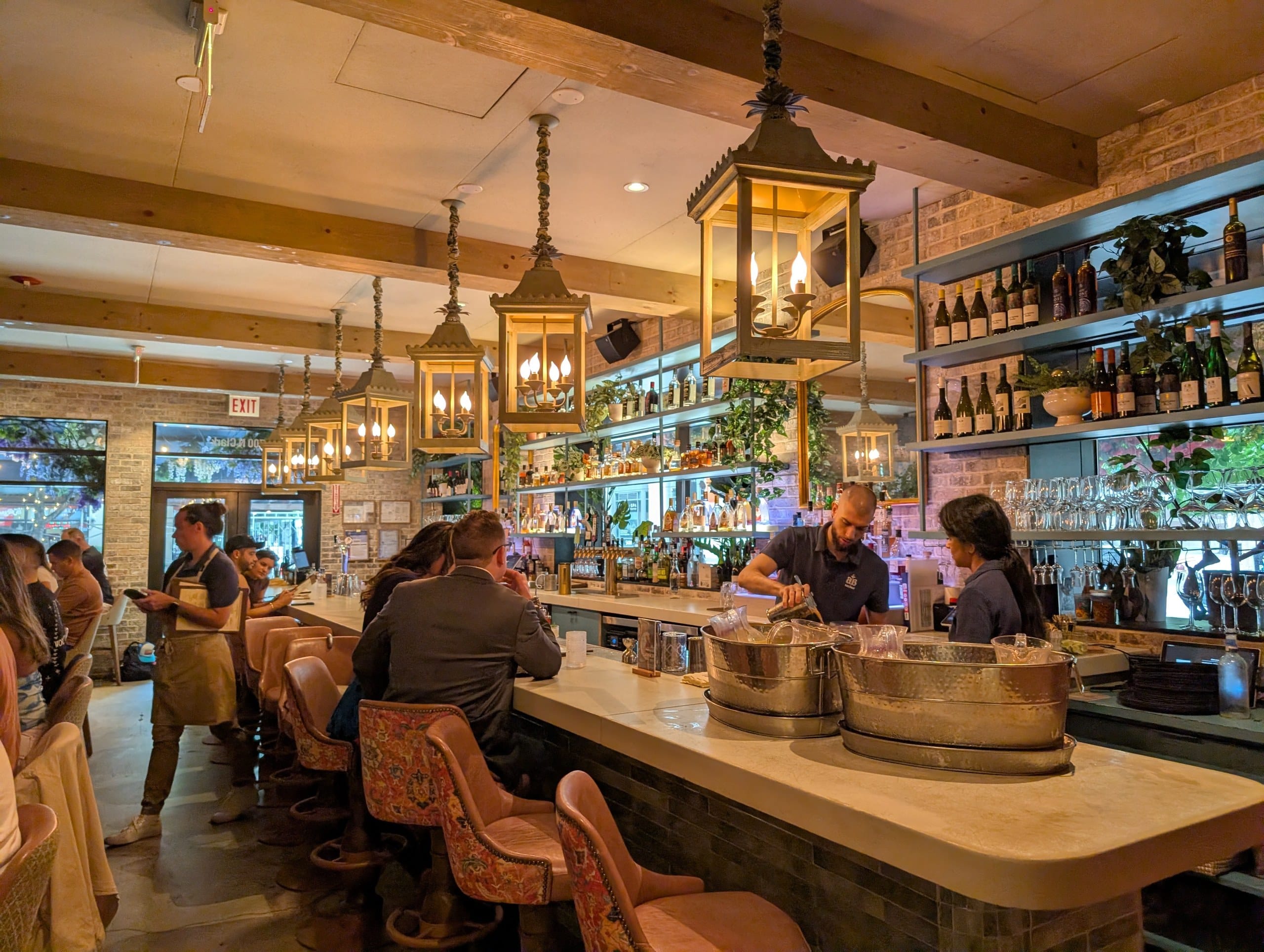 Iron lanterns hang the length of the marble bar, where bartenders work under shelves of wine and rakija against exposed brick.