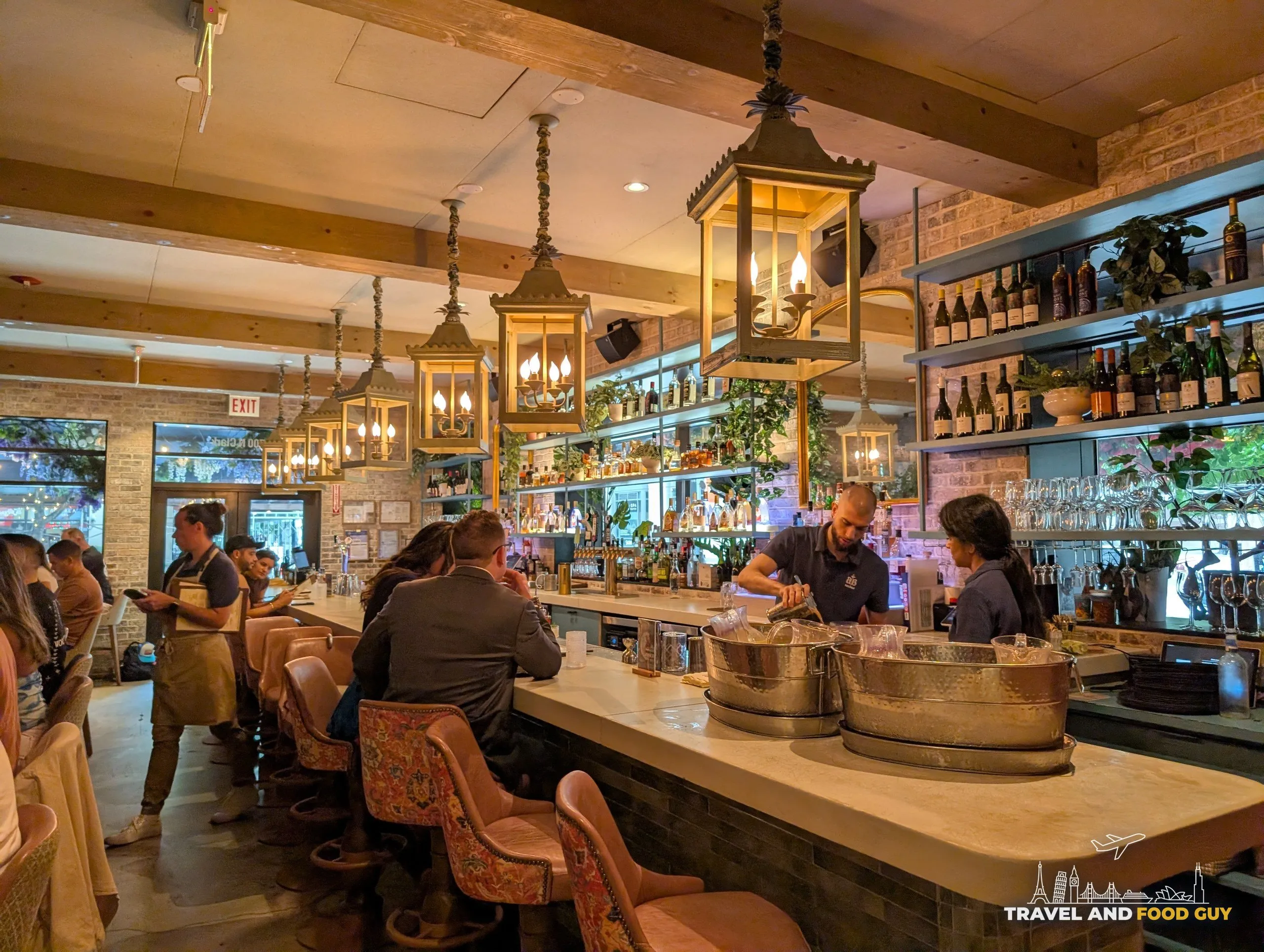 Iron lanterns hang the length of the marble bar, where bartenders work under shelves of wine and rakija against exposed brick.
