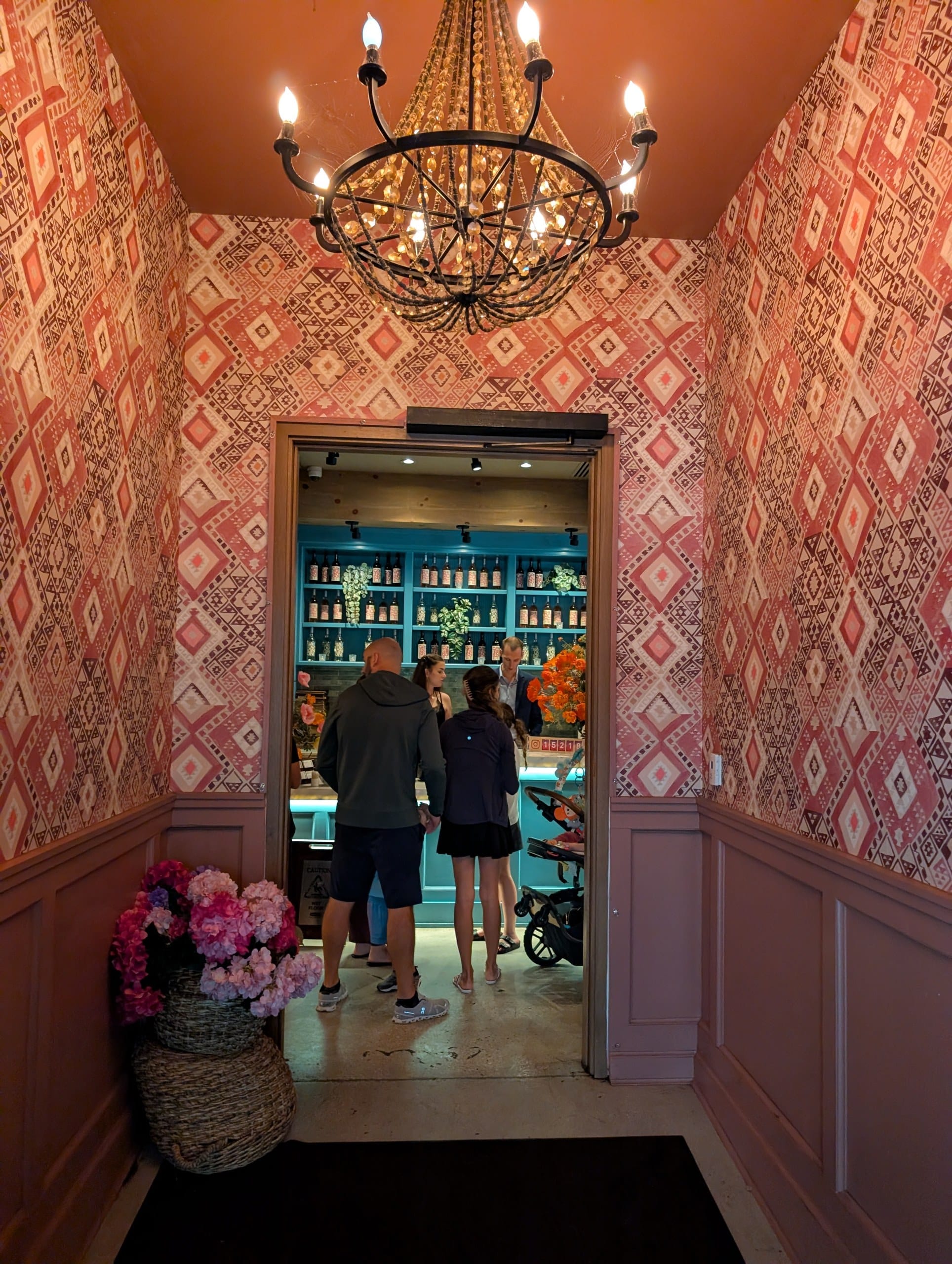Guests wait in a small vestibule wrapped in pink Balkan pattern wallpaper under a beaded chandelier, looking through to the host stand and bottle display.
