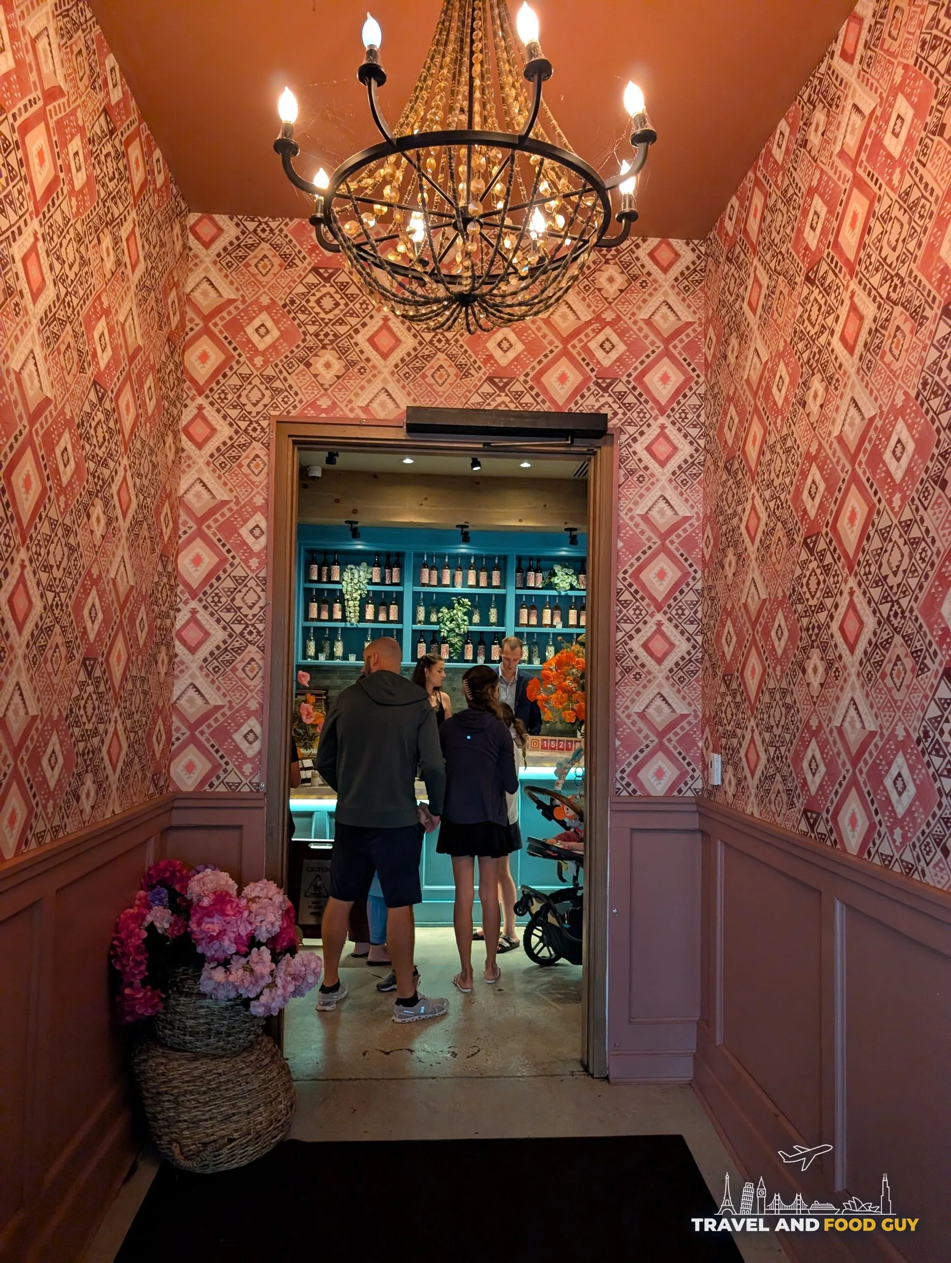 Guests wait in a small vestibule wrapped in pink Balkan pattern wallpaper under a beaded chandelier, looking through to the host stand and bottle display.