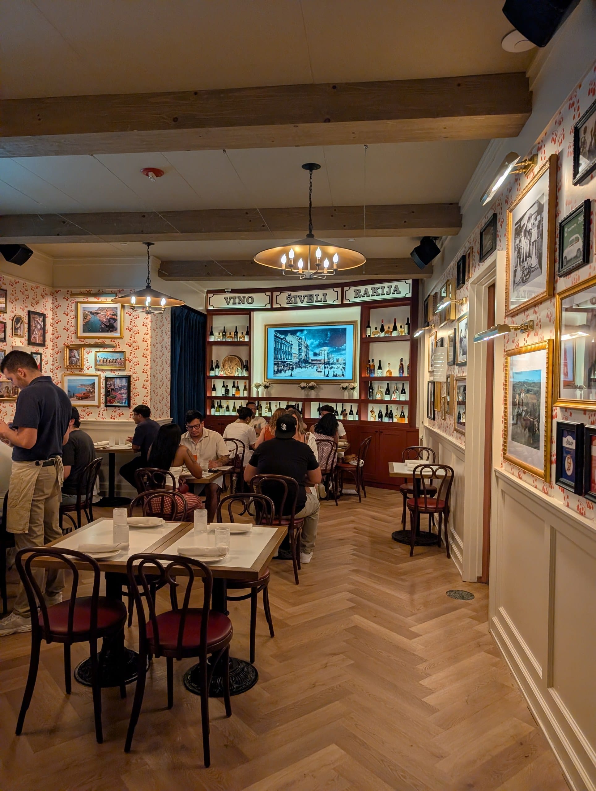 The front dining room is lined with gilt framed pictures and a back bar signed Vino, Ziveli and Rakija over bentwood chairs and herringbone floors.
