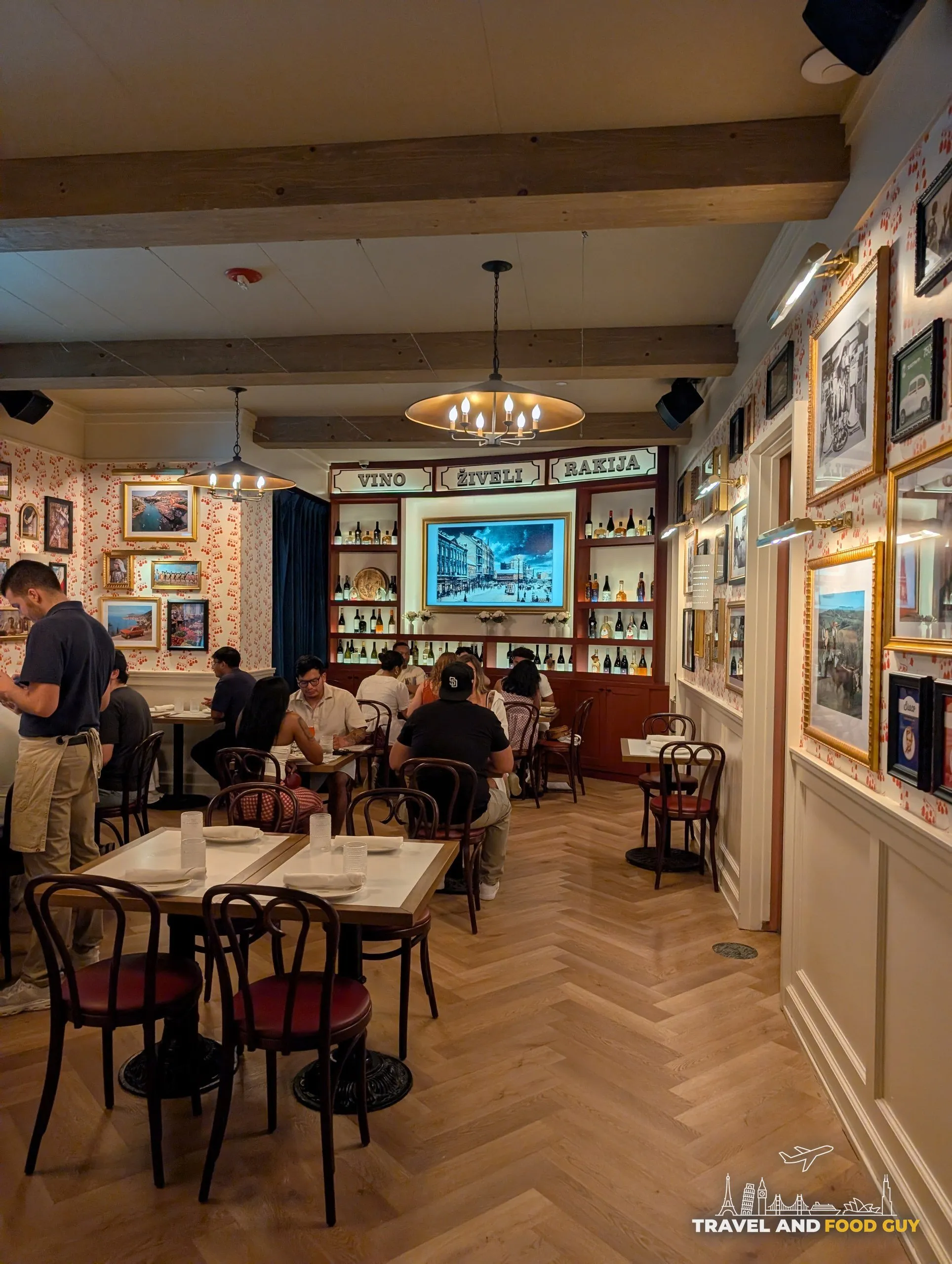 The front dining room is lined with gilt framed pictures and a back bar signed Vino, Ziveli and Rakija over bentwood chairs and herringbone floors.
