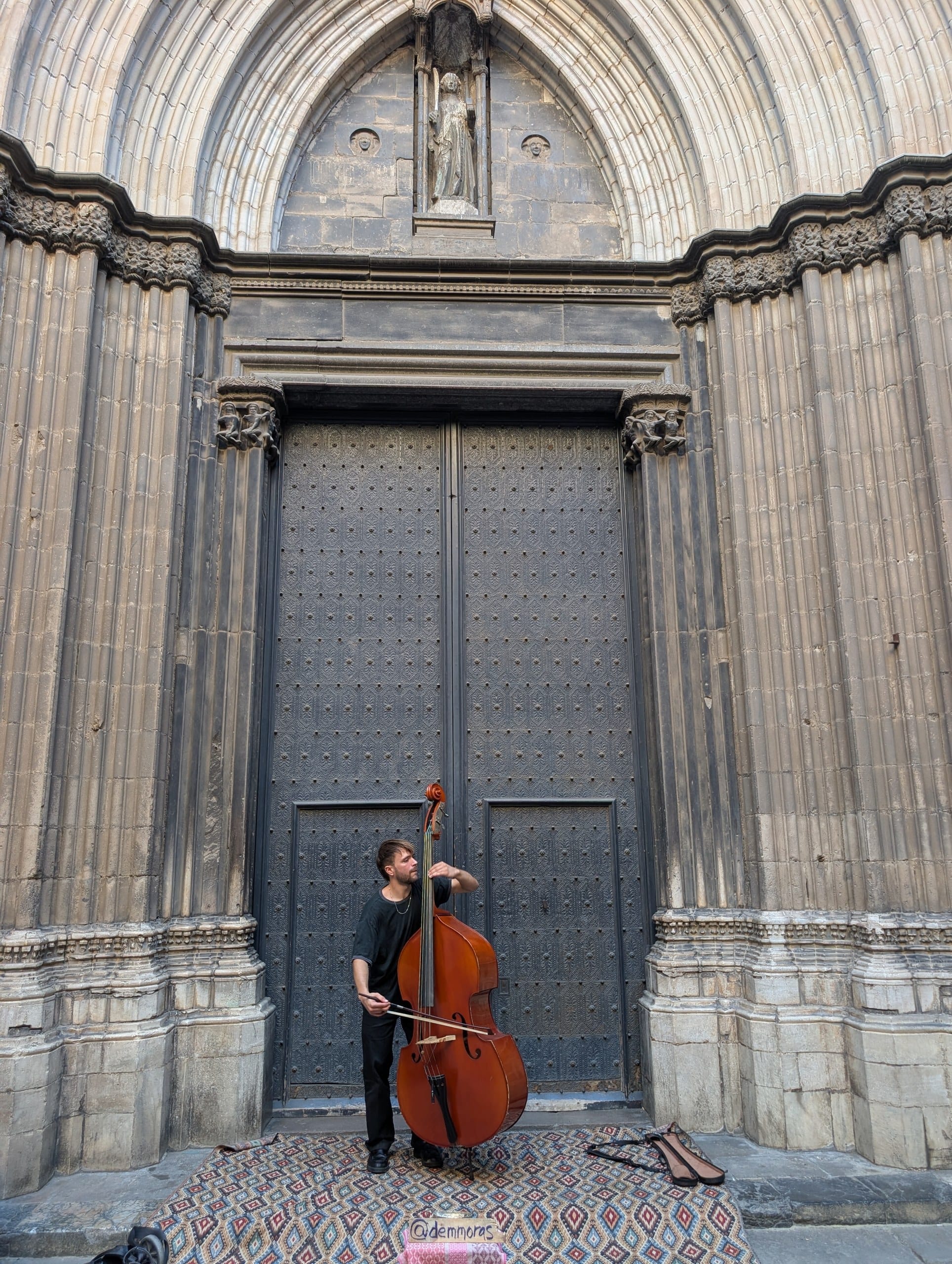 A street musician playing a double bass in front of the studded doors of Barcelona Cathedral, with a patterned rug and an open case on the ground