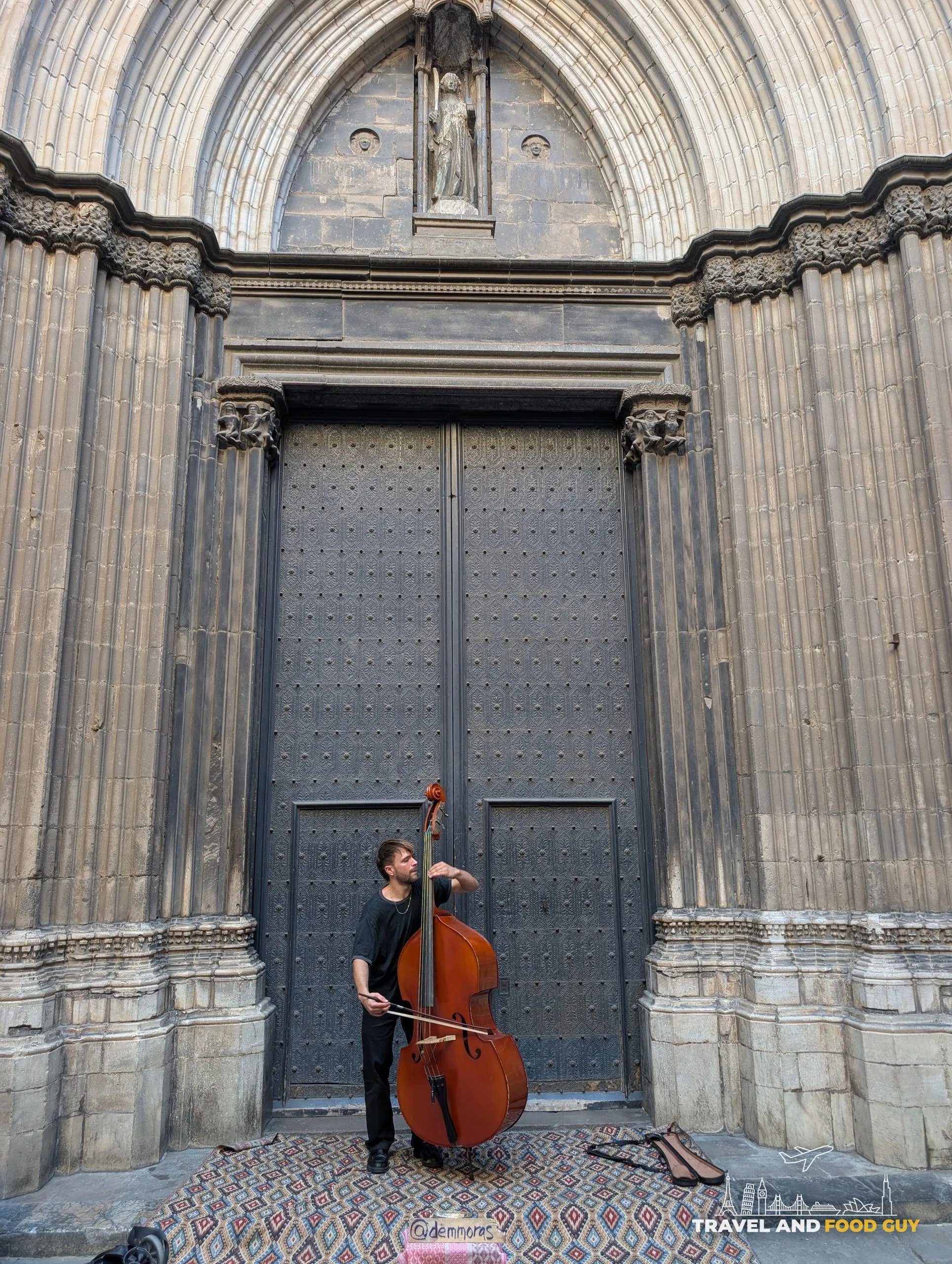 A street musician playing a double bass in front of the studded doors of Barcelona Cathedral, with a patterned rug and an open case on the ground