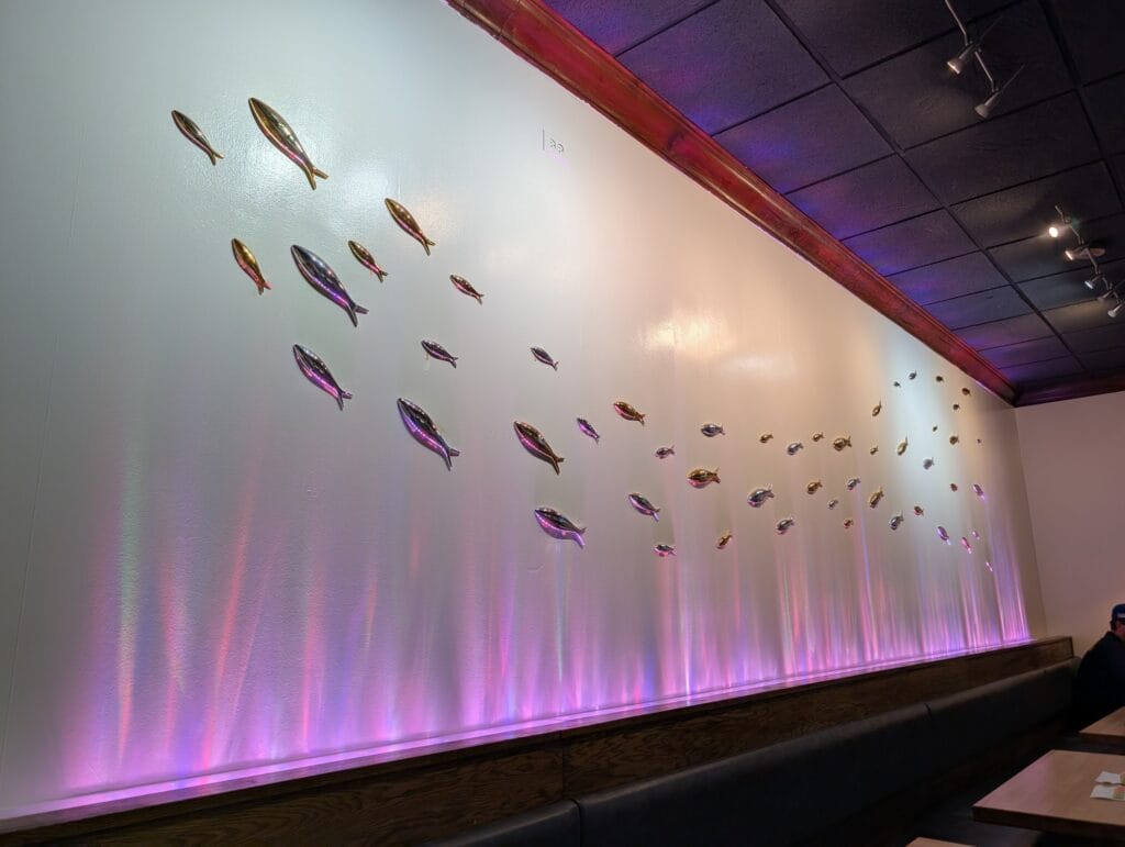 A shoal of polished metal fish running along the banquette wall at e Sushi, lit from below in shifting pink and violet.