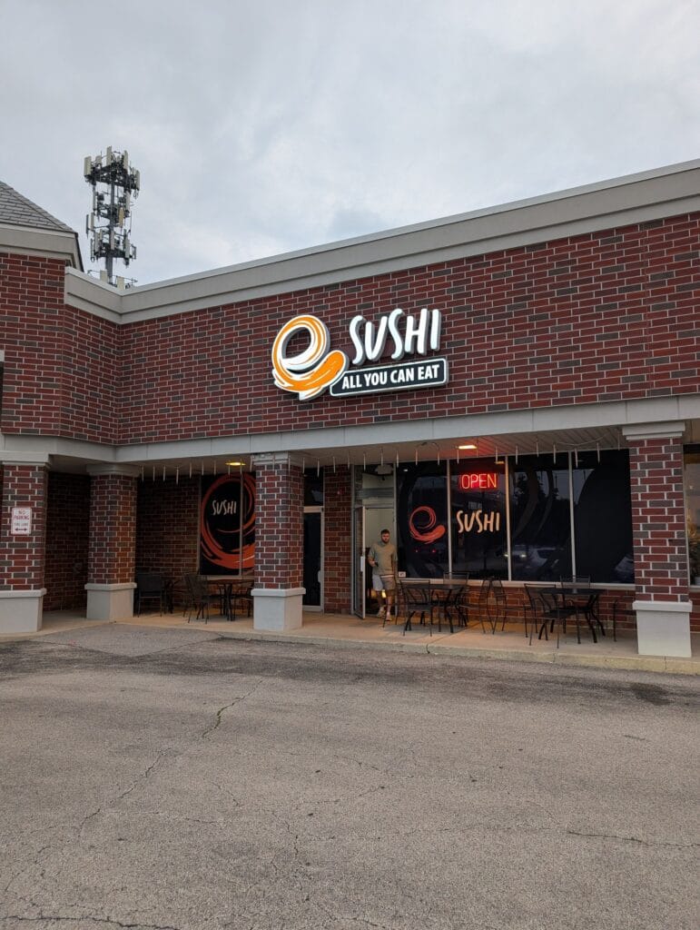 The entrance to e Sushi in Mount Prospect, with sidewalk tables and the neon OPEN sign in the window.