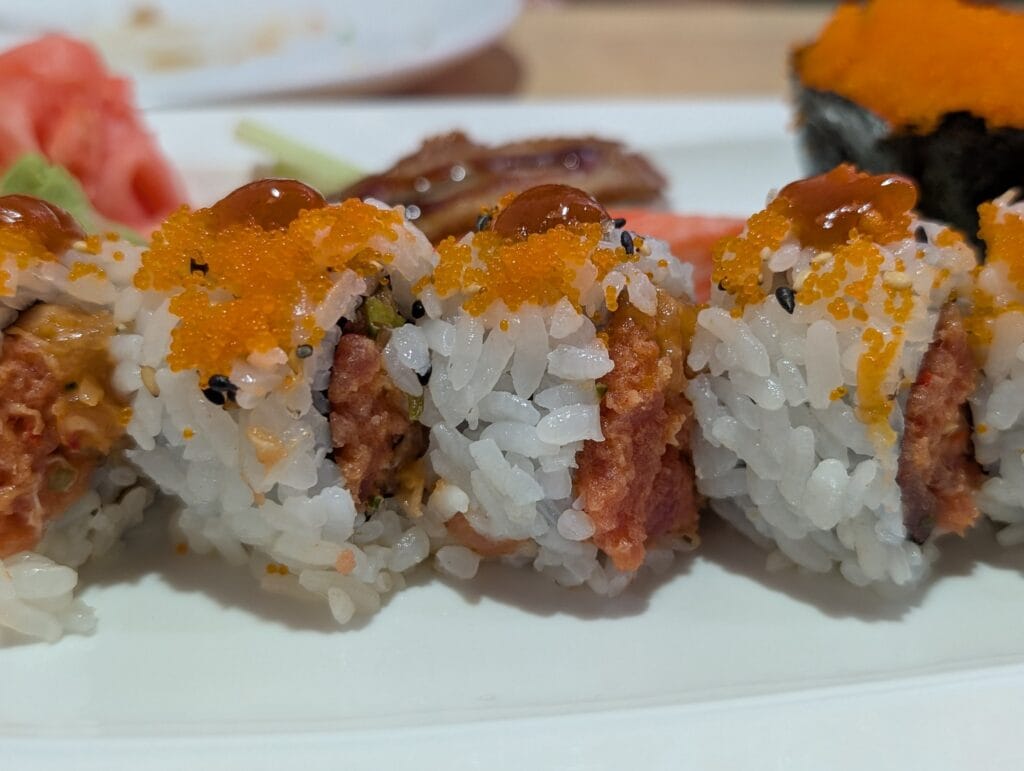 Close up of the Volcano roll at e Sushi, the spicy tuna coarse inside the rice, with orange tobiko, black and white sesame and a bead of chili sauce on each piece.