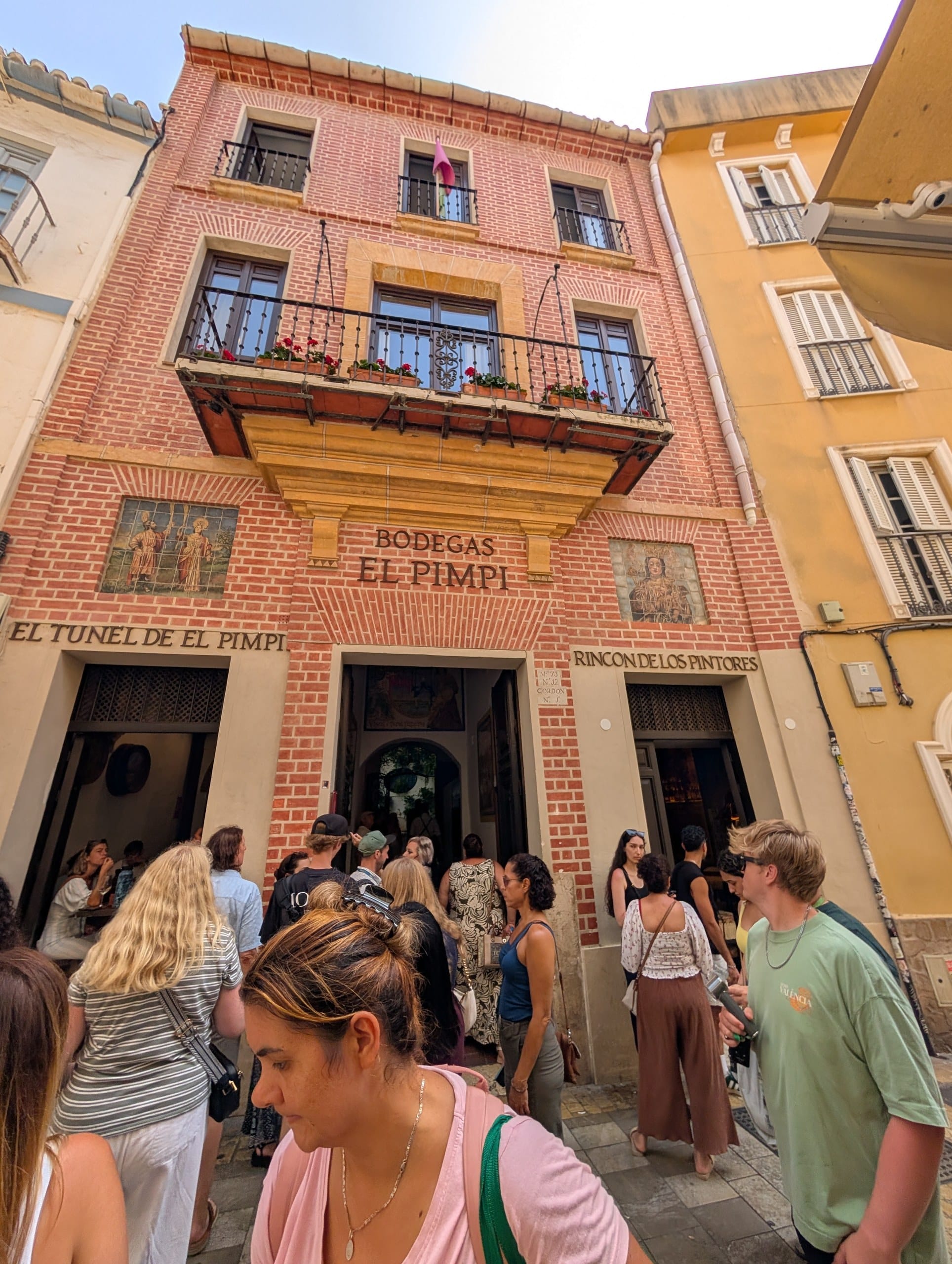Pink brick facade of Bodegas El Pimpi in Málaga with three named entrances and a crowd outside