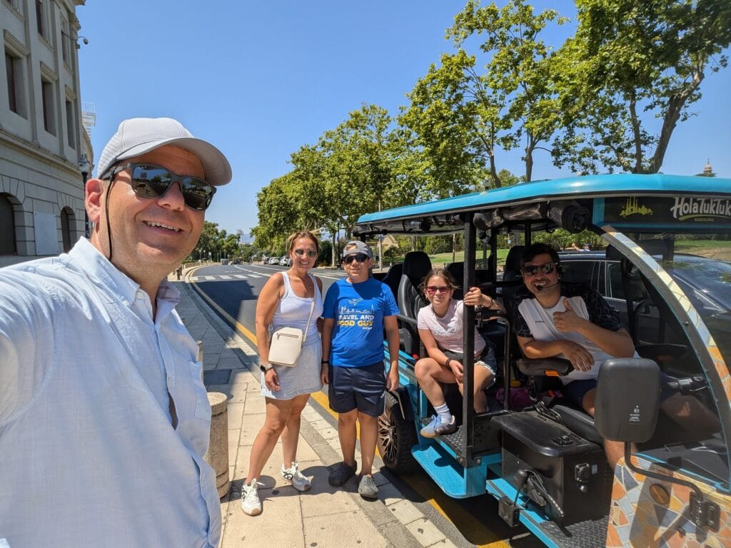 A family of four standing beside a blue Hola Tuk Tuk electric tuk tuk in Barcelona with their guide Leo at the wheel
