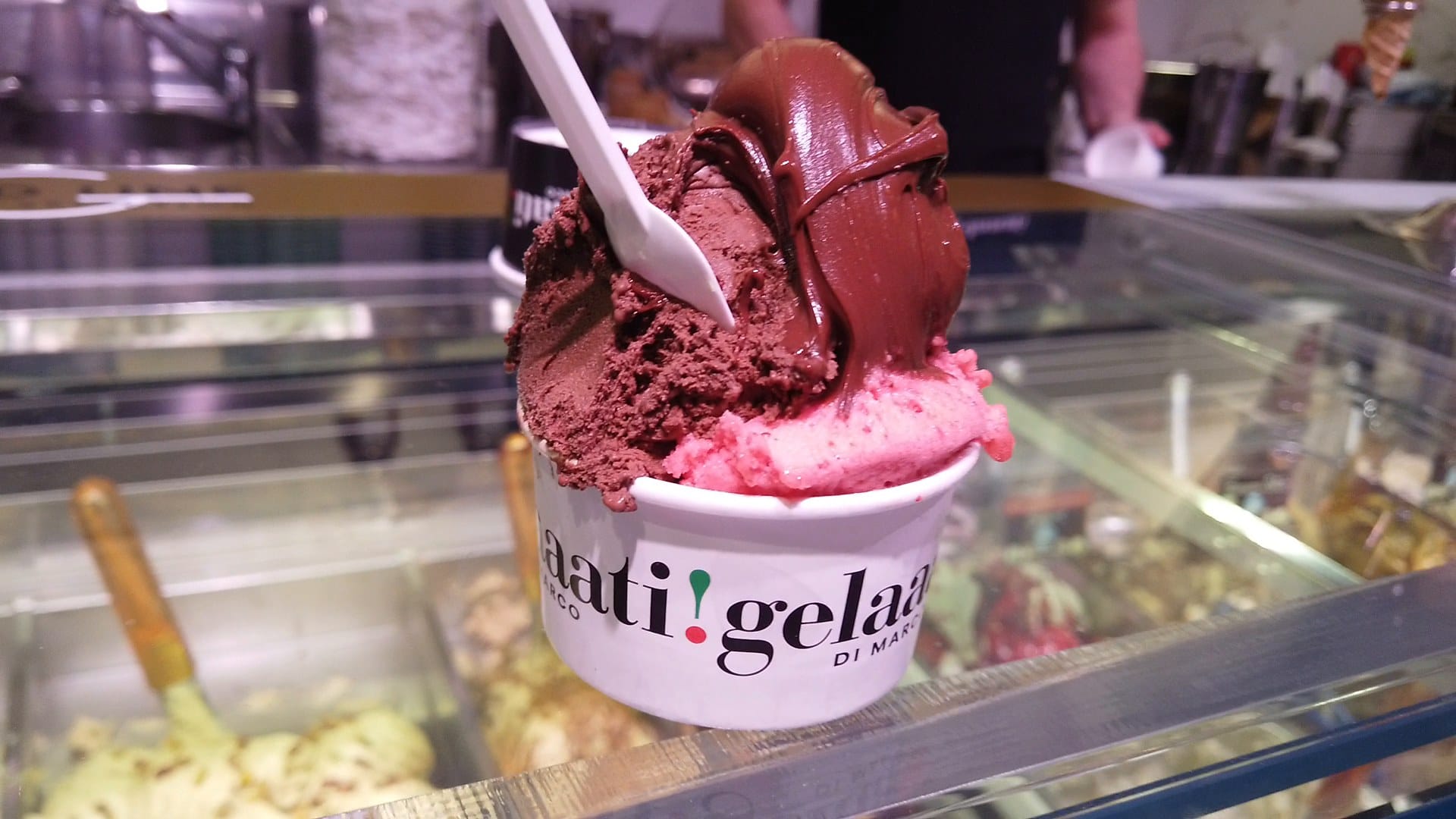 A branded cup of chocolate gelato with chocolate sauce over a bright raspberry scoop, held above the gelato case at Gelaaati DI MARCO in Barcelona