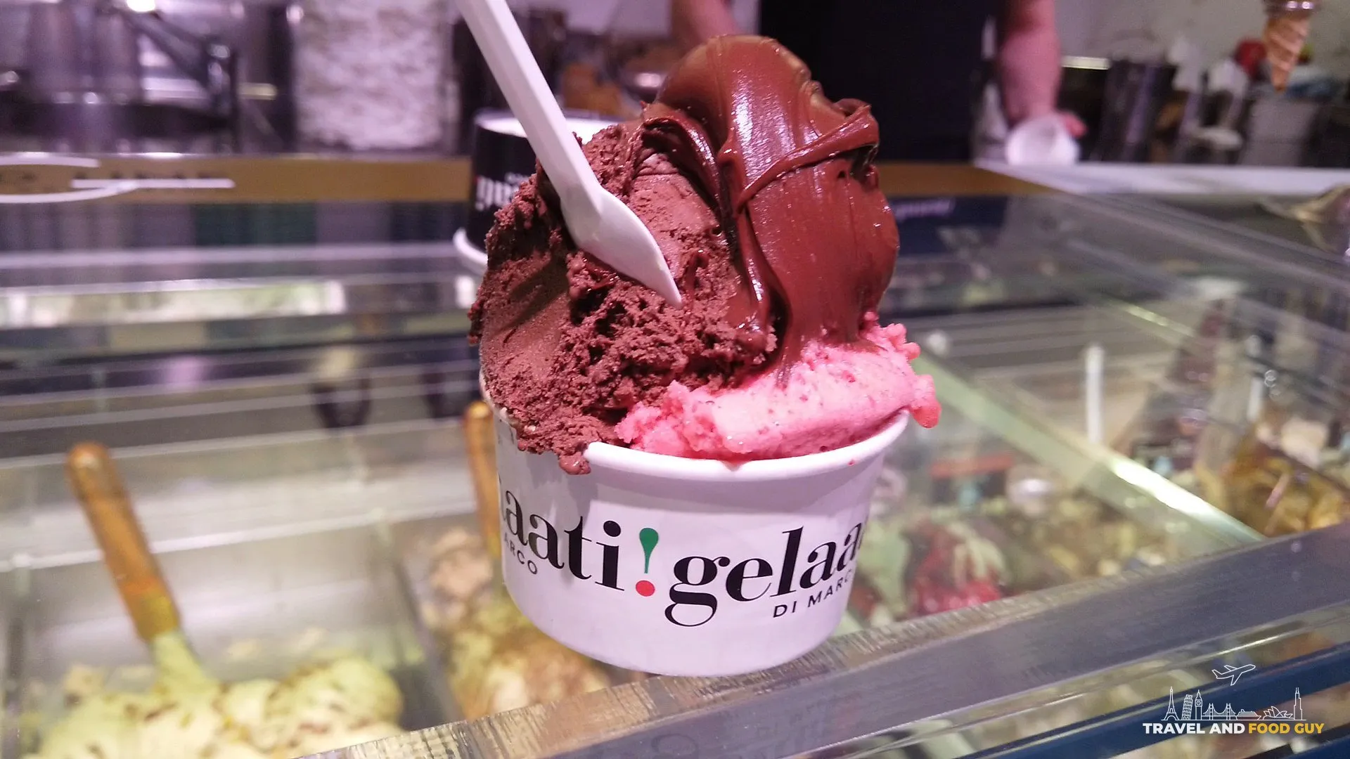 A branded cup of chocolate gelato with chocolate sauce over a bright raspberry scoop, held above the gelato case at Gelaaati DI MARCO in Barcelona