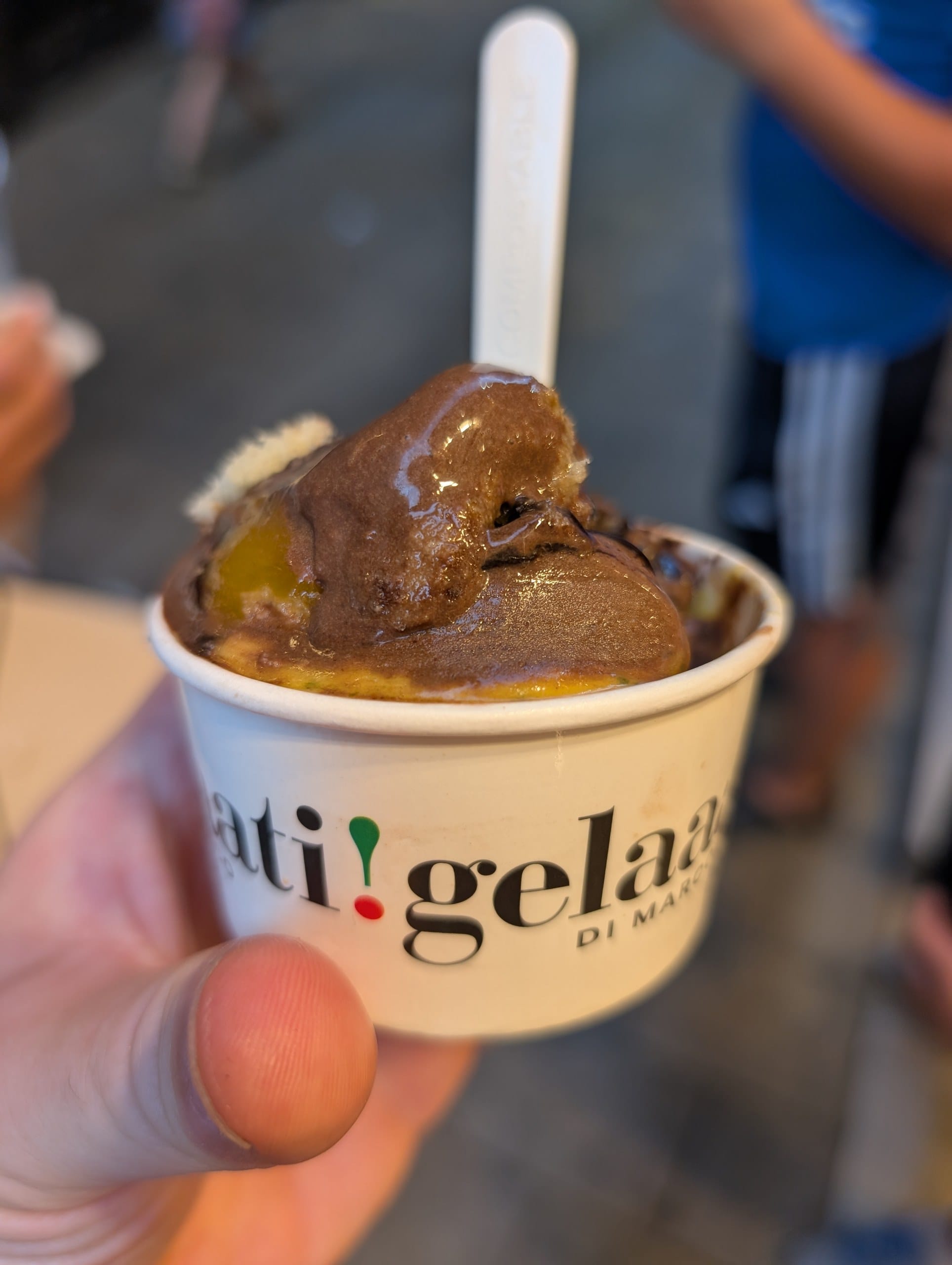 A branded cup of gelato with a chunky brown scoop over a peach scoop, held up outside Gelaaati DI MARCO in Barcelona