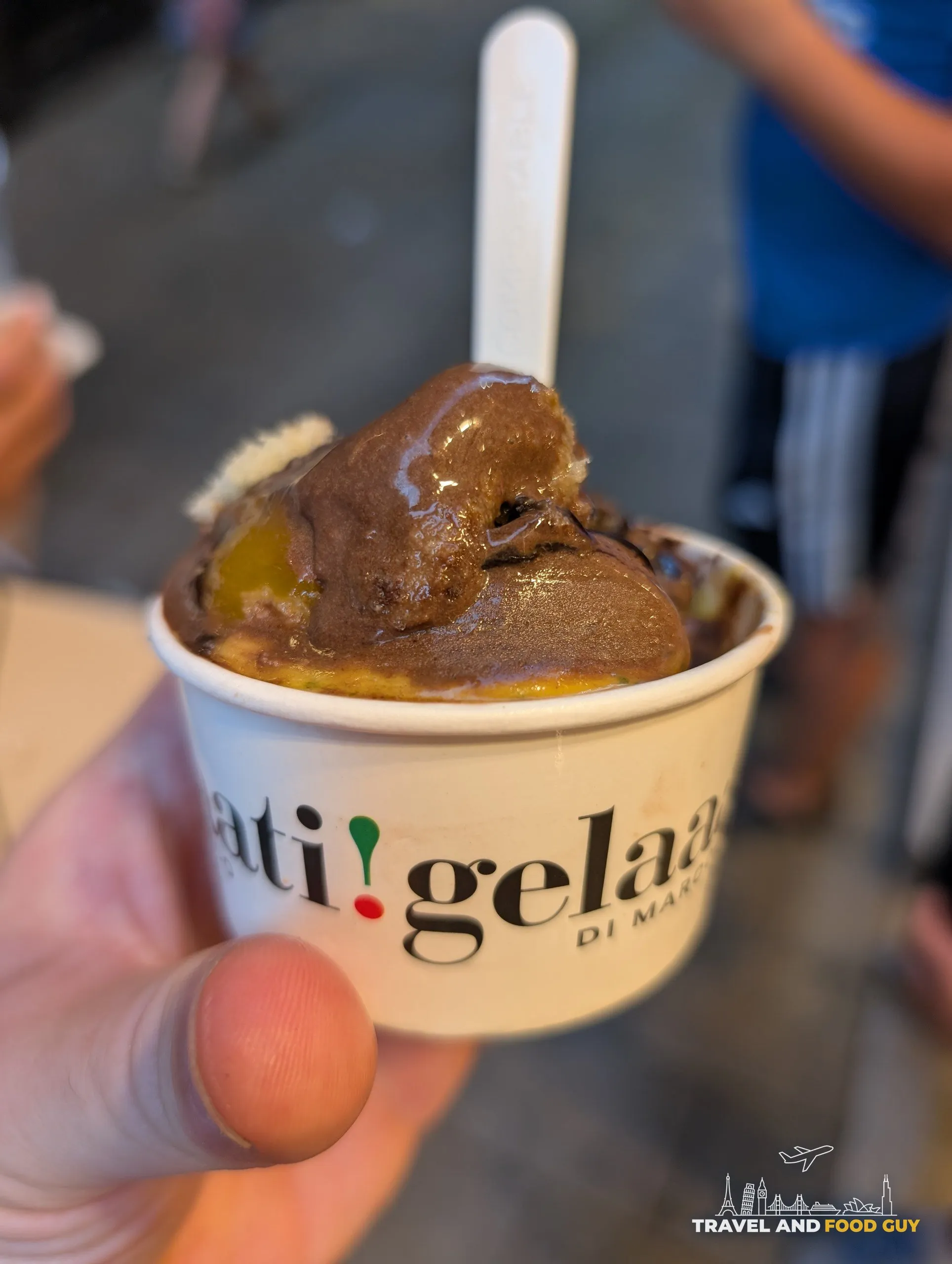 A branded cup of gelato with a chunky brown scoop over a peach scoop, held up outside Gelaaati DI MARCO in Barcelona