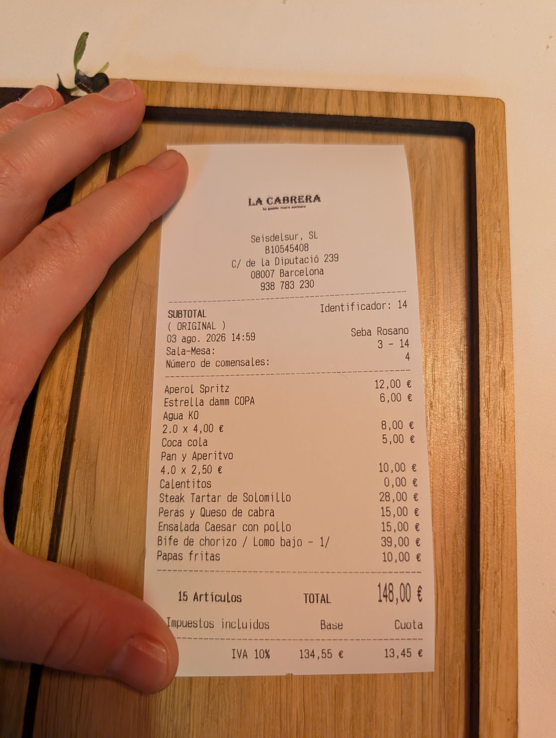 The itemized La Cabrera Barcelona bill for four people showing Aperol Spritz 12,00, Estrella Damm 6,00, two waters at 4,00, Coca-Cola 5,00, pan y aperitivo four at 2,50, calentitos 0,00, steak tartar de solomillo 28,00, peras y queso de cabra 15,00, ensalada Caesar con pollo 15,00, bife de chorizo lomo bajo half portion 39,00 and papas fritas 10,00, totaling 148,00 EUR including 10 percent IVA