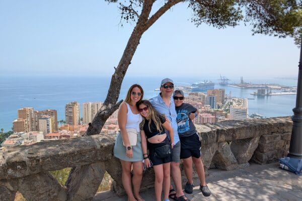 The four of us at a stone wall on a hilltop viewpoint above Malaga, with the city, the beach high rises, the container cranes and a cruise ship at the dock below.