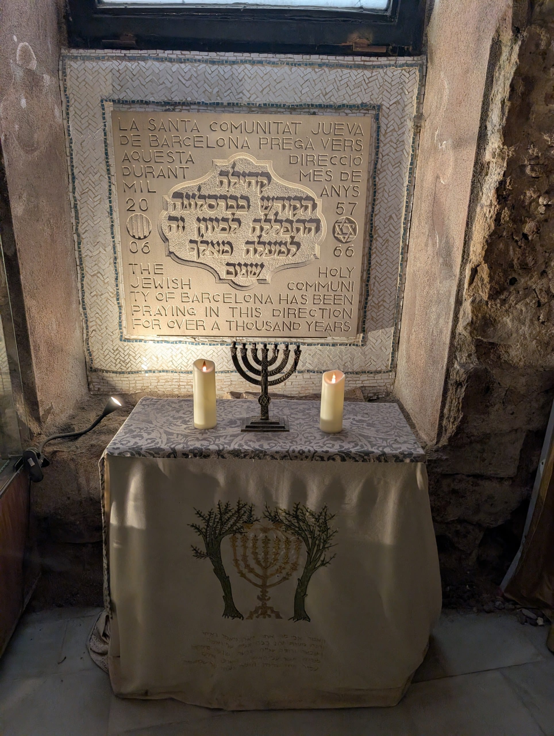 A stone memorial tablet in Hebrew and English above a draped table holding a menorah and two lit candles at the Sinagoga Major in Barcelona