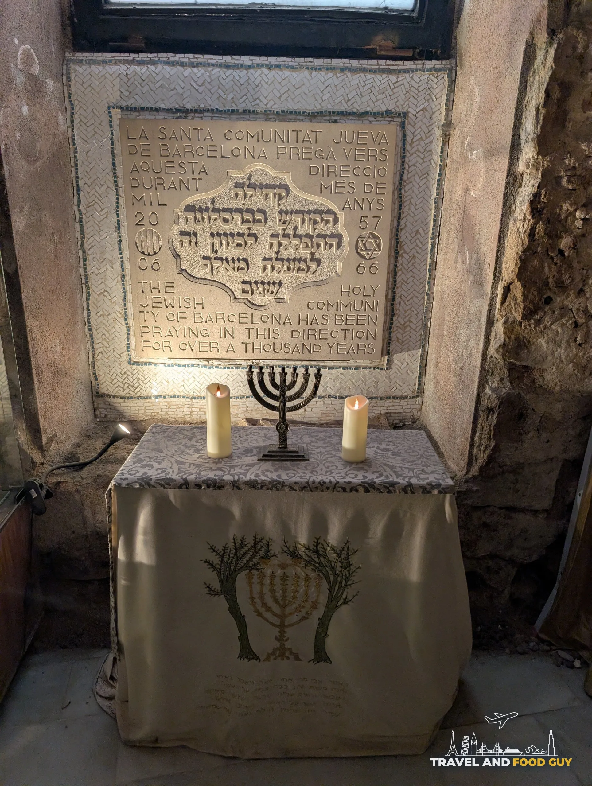 A stone memorial tablet in Hebrew and English above a draped table holding a menorah and two lit candles at the Sinagoga Major in Barcelona