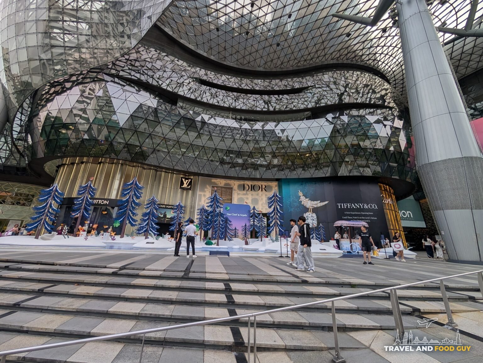 The glass canopy of ION Orchard mall on Orchard Road with a blue Christmas tree installation and Louis Vuitton, Dior, and Tiffany storefronts visible