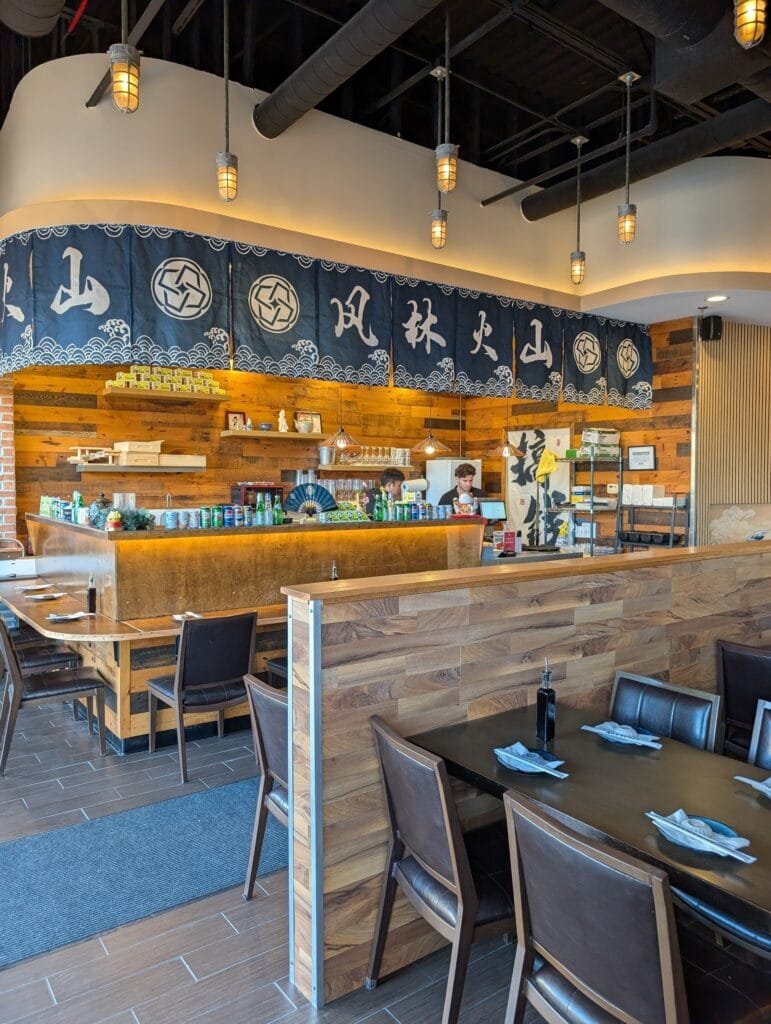 The sushi counter under a navy noren printed with the four Furinkazan characters, tables in the foreground.