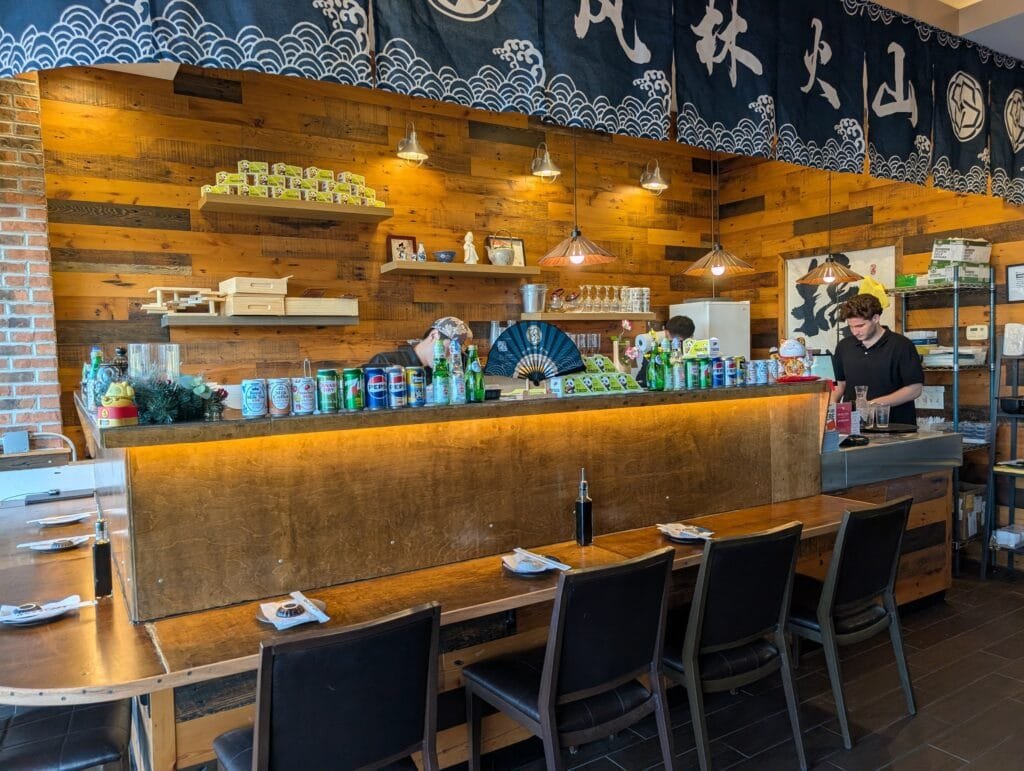 The long wooden sushi counter with stools, drink cans lined along the top and staff working behind it.