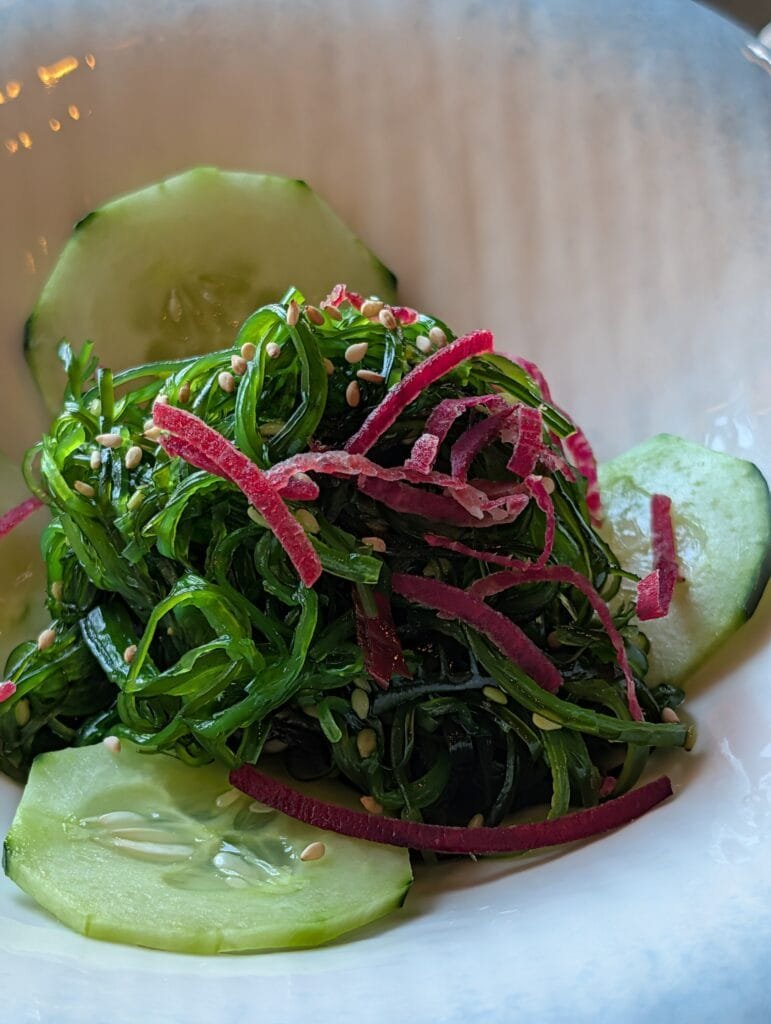Seaweed salad with sesame and shredded pickled radish over cucumber slices in a white bowl.