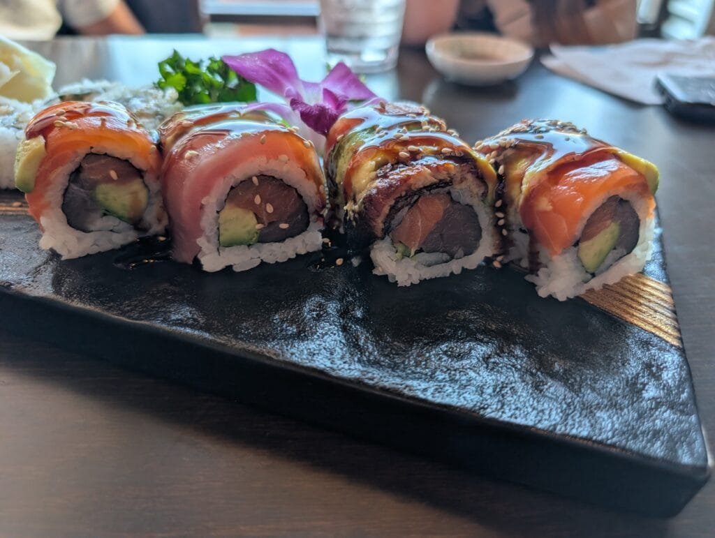 The crouching tiger roll on a black board, topped with salmon, tuna and avocado under eel sauce.