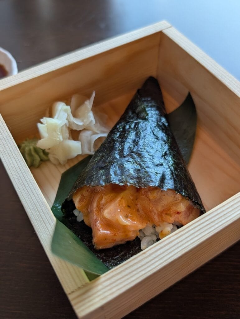 A spicy salmon hand roll standing in a small wooden box with pickled ginger and wasabi.