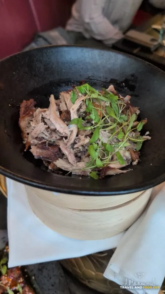 Shredded aromatic crispy duck topped with micro herbs in a black bowl over bamboo steamers at The Ivy Asia St Paul's.