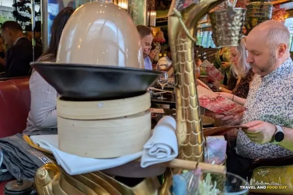 The Aromatic Half Crispy Duck arriving at The Ivy Asia St Paul's under a glass dome on a gold bird-shaped stand, cherry blossoms overhead.