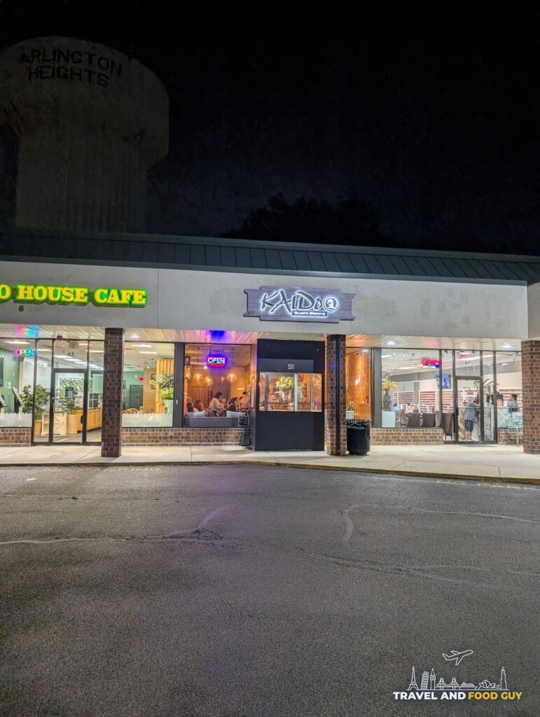 The Kaido Sushi Bistro storefront at night in its Golf Road strip mall in Arlington Heights, with the water tower behind it.