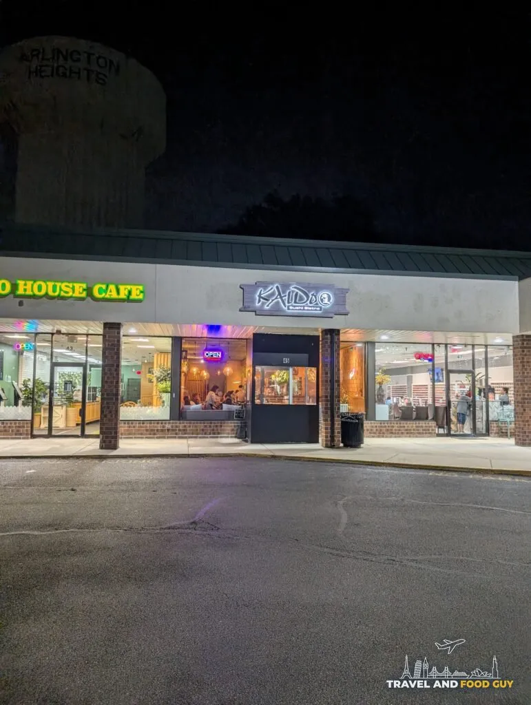 The Kaido Sushi Bistro storefront at night in its Golf Road strip mall in Arlington Heights, with the water tower behind it.