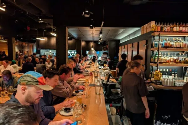 The long wood sushi bar at Sushi-San in River North, full of diners, with the bartenders and the bottle wall on the right.