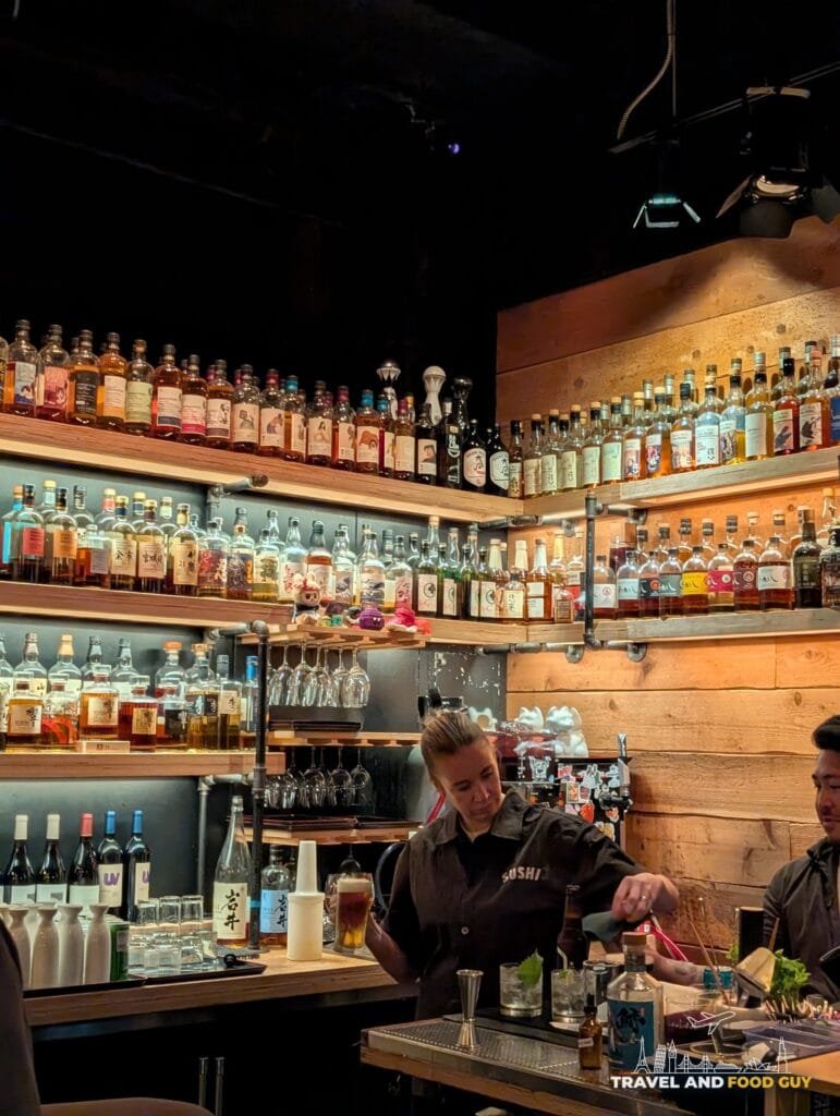 Shelves of Japanese whisky bottles stacked to the ceiling behind the bar at Sushi-San, a bartender pouring a beer.