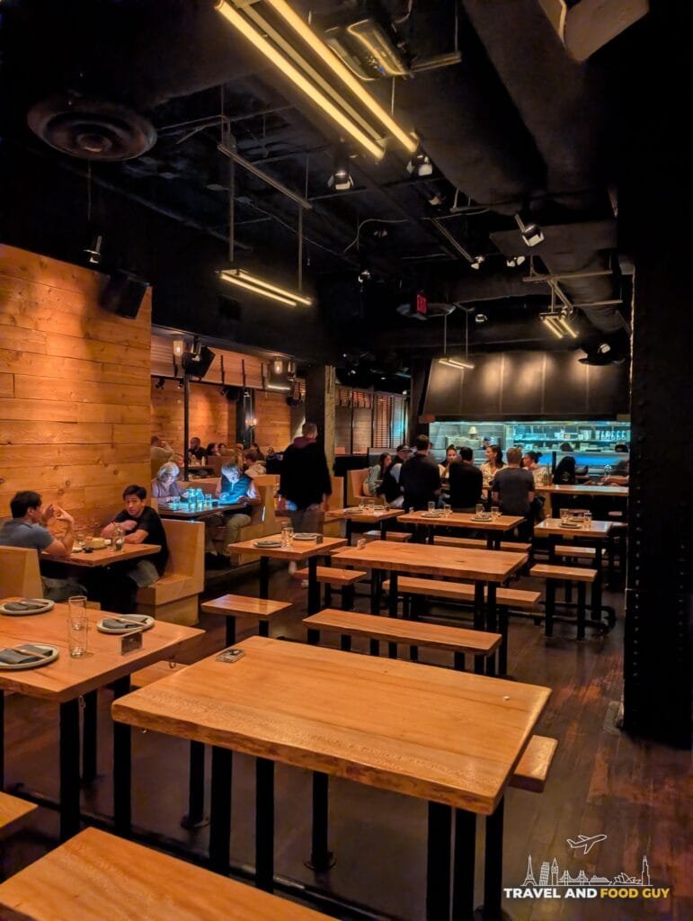 Sushi-San's main dining room with live edge wood tables and benches, the open kitchen glowing at the back.