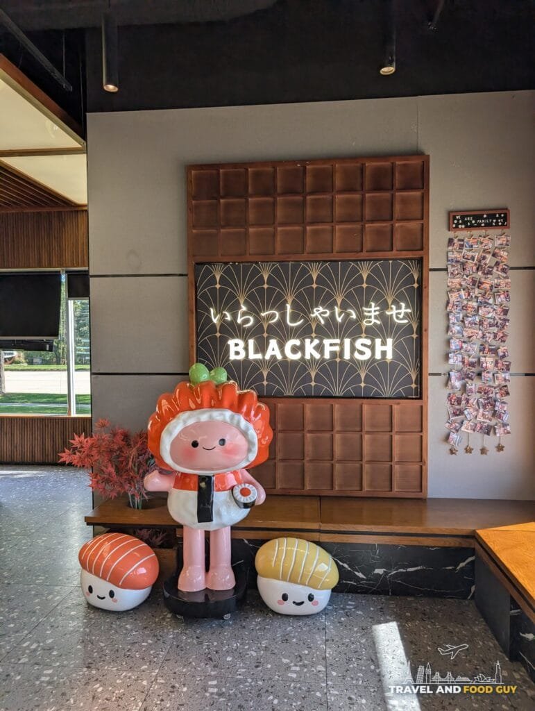 A Blackfish welcome sign in Japanese and English with a salmon nigiri mascot statue and two sushi plushies.