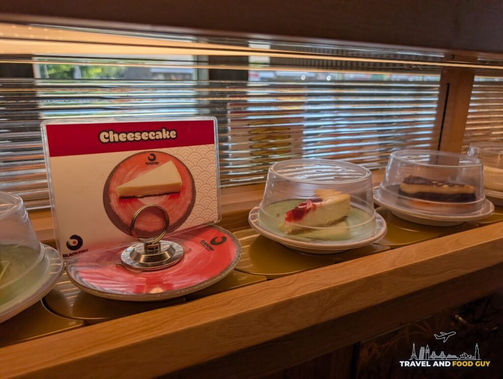 A slice of cheesecake under a clear dome on the conveyor belt with a picture card behind it.