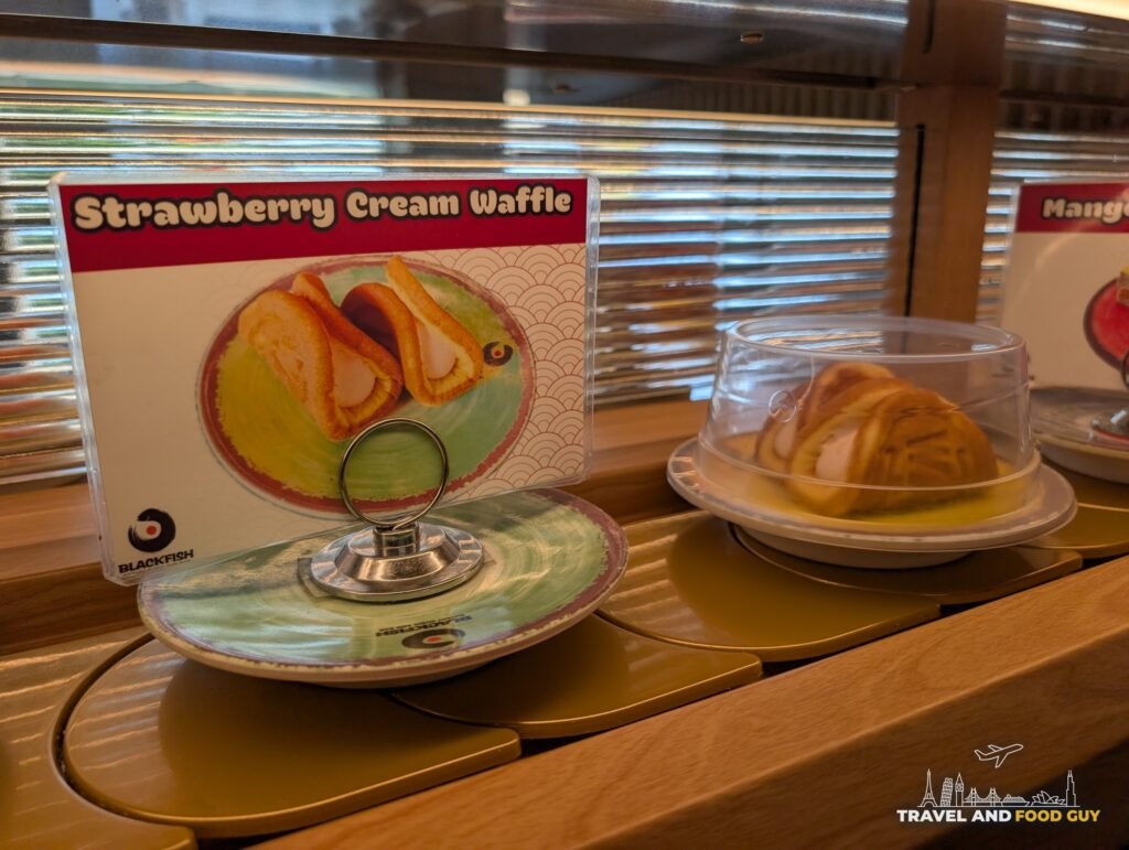 A strawberry cream waffle under a clear dome on the conveyor belt at Blackfish with its picture card.