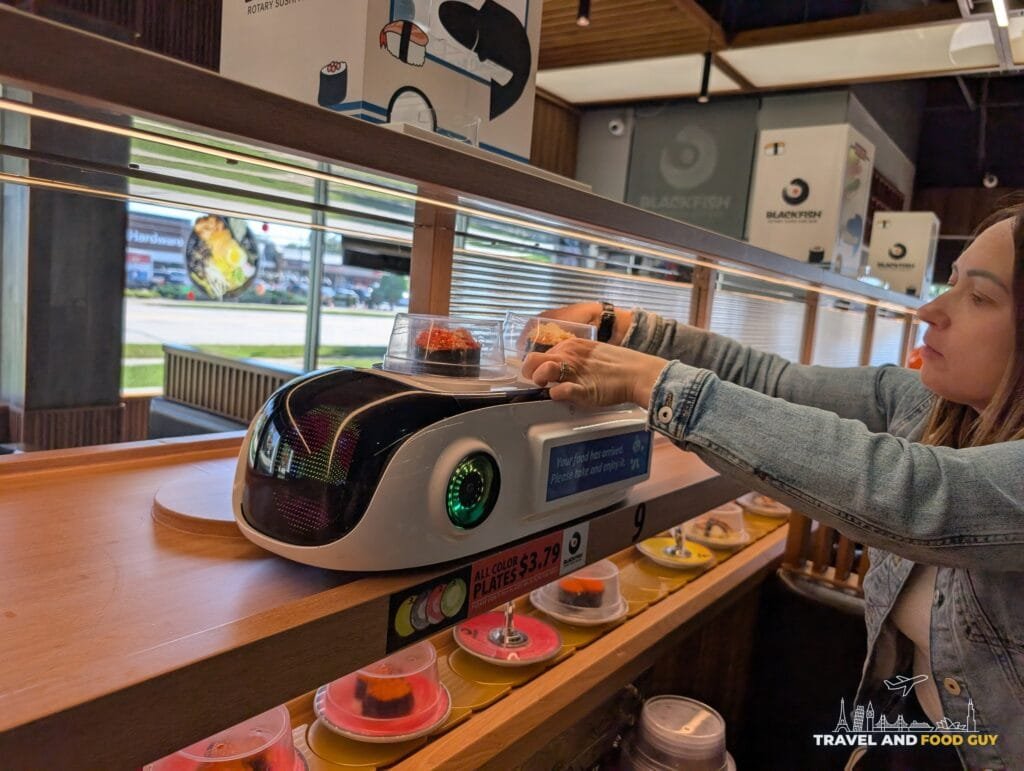 Vika reaching up to take two plates of gunkan off the robot train that runs on a track above the conveyor belt at Blackfish.