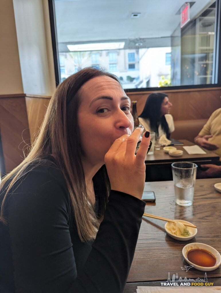 Vika eating a hand roll at a table in the SUGARFISH Beverly Hills dining room, with wood panels and a big window behind her.